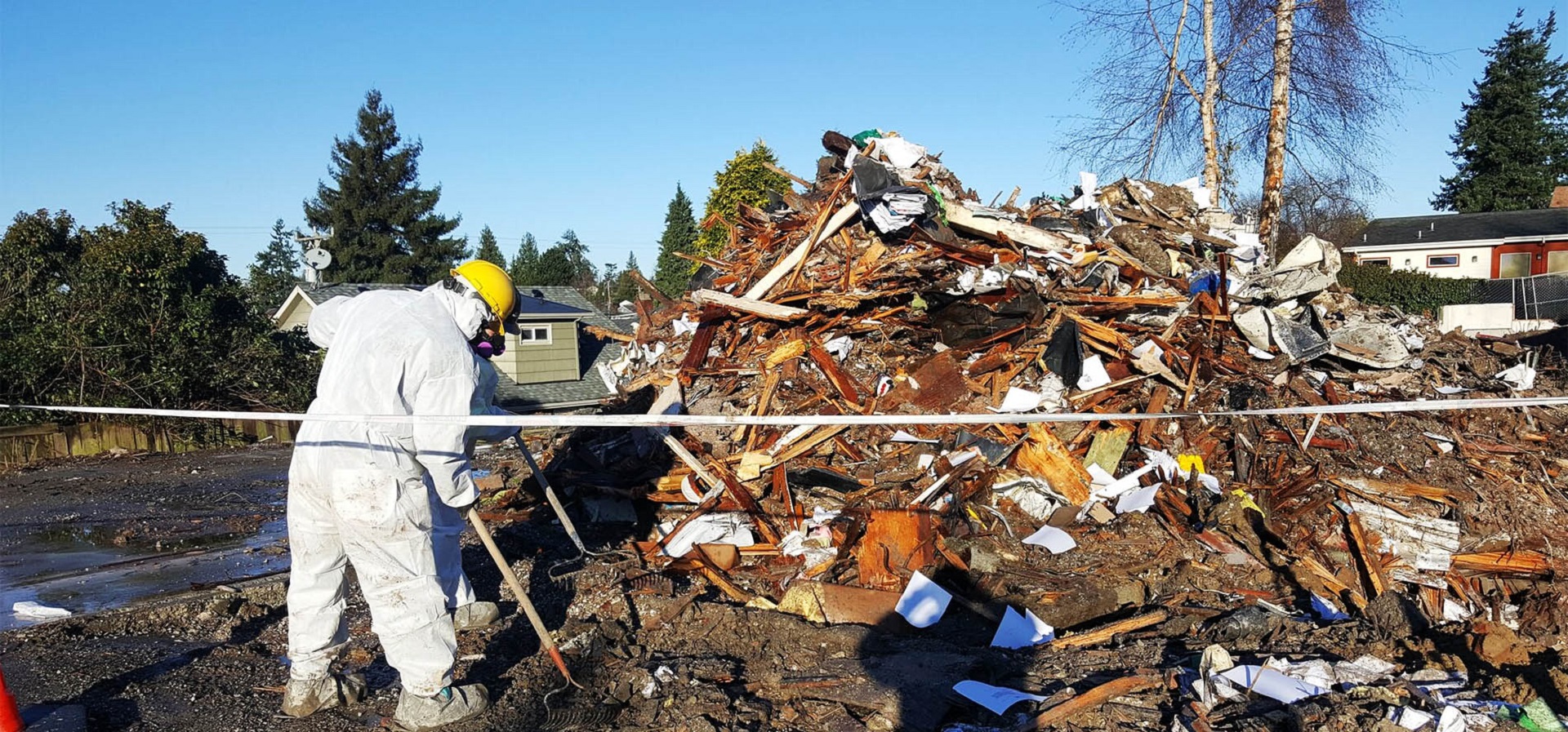 Two professional demolition contractors in white coveralls and PPE covering their heads raking the debris from a demolition project into a pile.