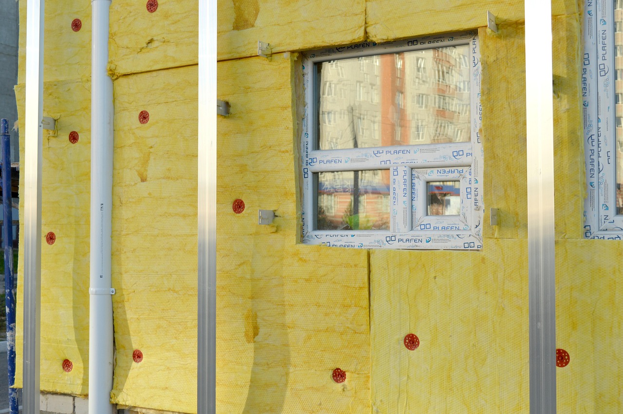 The exposed siding of a building showing insulation material beneath PVC piping, support beams, and around a window.